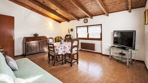 Ein gemütliches Wohnzimmer mit Holzbalkendecke, einem Essbereich mit Tisch und Stühlen, einer Couch, einem Fernseher auf einem Schrank und einem Fenster mit Vorhängen.