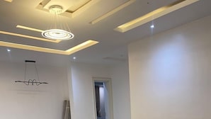 Ein modernes Wohnzimmer mit einem großen, winkelförmigen Sofasessel, einem gemusterten Teppich und einer auffälligen Deckenleuchte.