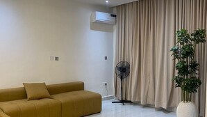 Ein modernes Wohnzimmer mit einem Ecksofa, einem Standventilator, einer Topfpflanze und einem großen Fenster mit Vorhängen.