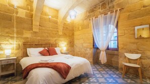 A bedroom with a stone wall, a bed with white bedding and red pillows, a wooden nightstand with a lamp, a chair, and a window with a sheer curtain. - San Lawrenz Farmhouse Rustic Retreat With Pool (San Lawrenz)