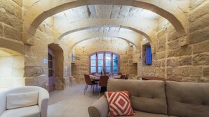 A stone-walled room with a dining area, a sofa with a patterned pillow, and a view through a window. - San Lawrenz Farmhouse Rustic Retreat With Pool (San Lawrenz)