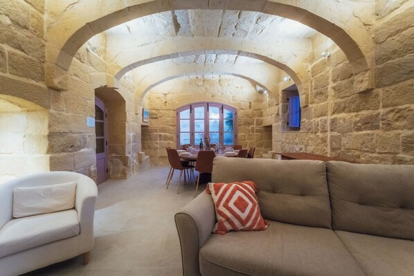 A stone-walled room with a dining area, a sofa with a patterned pillow, and a view through a window. - San Lawrenz Farmhouse Rustic Retreat With Pool (San Lawrenz)