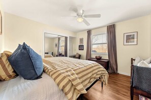 Ein Schlafzimmer mit einem Bett, einem Nachttisch, einem Spiegel, einem Fenster mit Vorhängen und einem Deckenventilator.