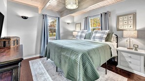A bedroom with a large bed, a nightstand, a dresser, and a ceiling fan. There are windows with curtains, a wall-mounted lamp, and a framed picture on the wall. - The Oak Cottage- Charming Downtown Retreat in the Heart of Siloam Springs (Siloam Springs)