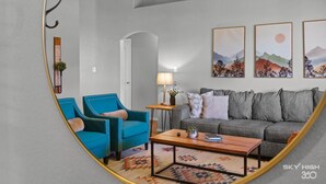 Una sala de estar con un sillón azul, un sofá gris, una mesa de centro de madera y tres obras de arte enmarcadas en la pared.