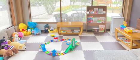 A cozy room with a large window, a sofa, a wooden table, and a bookshelf filled with children's books.