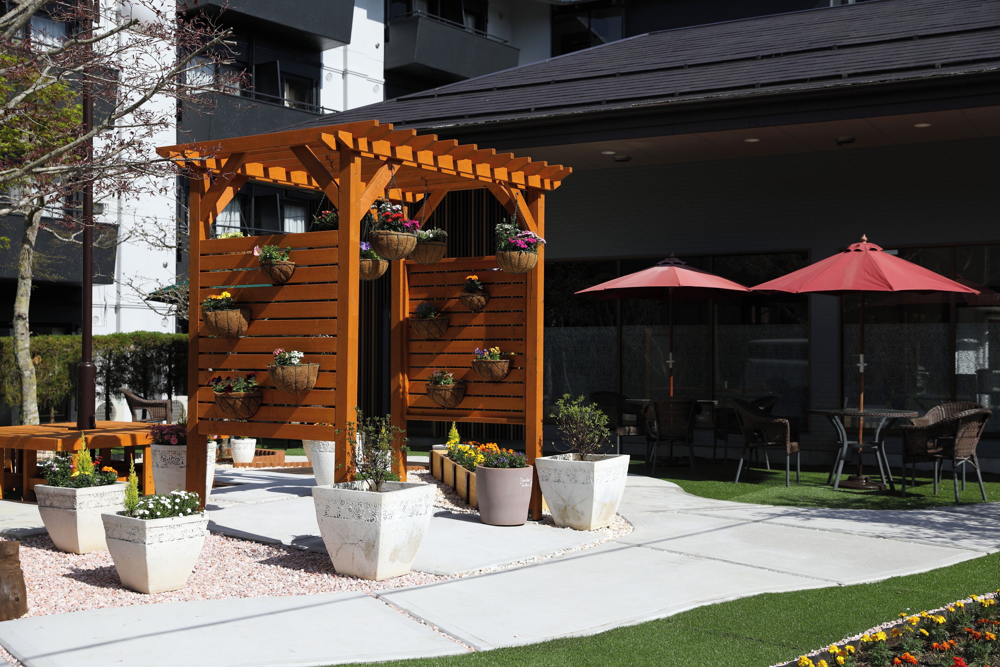 Una pérgola de madera con cestas de flores colgantes, rodeada de plantas en macetas y un patio con mobiliario exterior.
