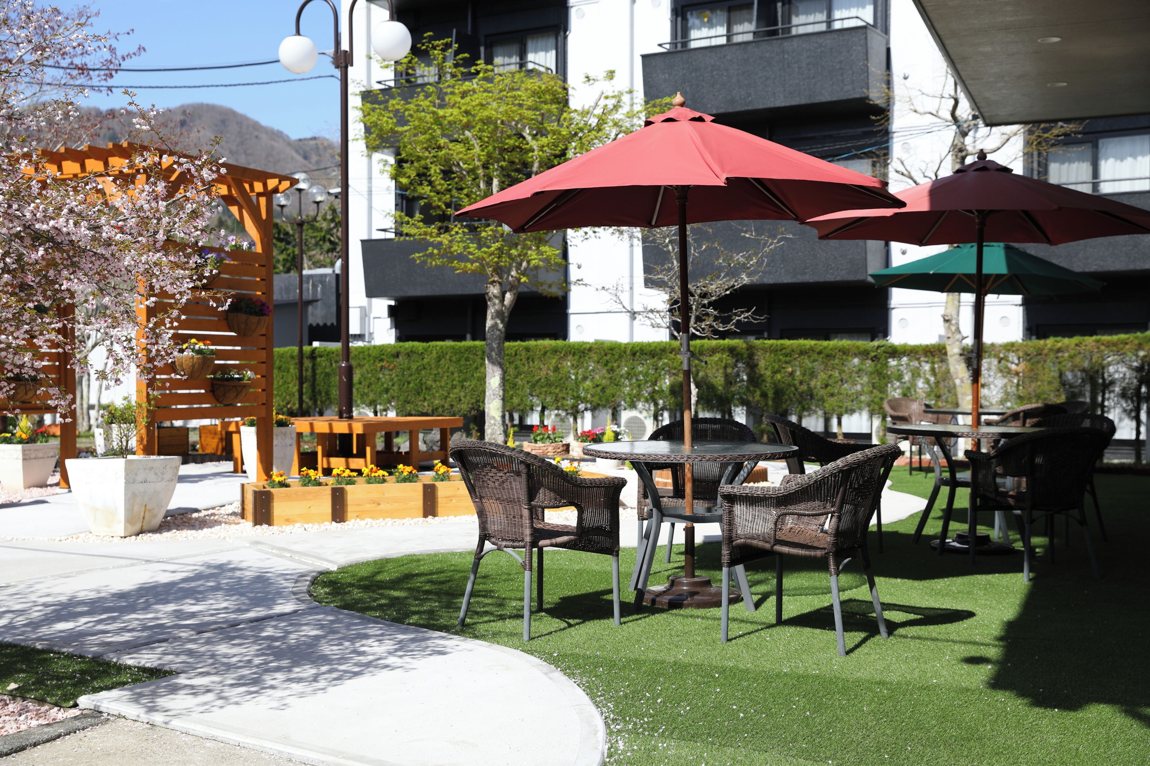 An outdoor patio area with wicker furniture, red umbrellas, and a wooden pergola with flowering plants.