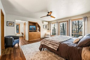 Ein Schlafzimmer mit Bett, Deckenventilator, Fernseher, Kleiderschrank, Stuhl und großen Fenstern.