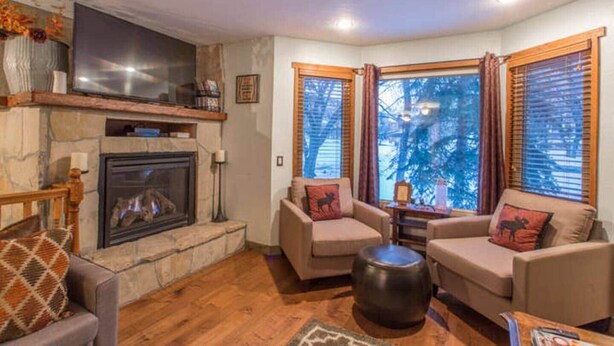 Ein gemütliches Wohnzimmer mit Kamin aus Stein, zwei Sofas mit dekorativen Kissen, einem Couchtisch und einem großen Fenster mit Vorhängen.