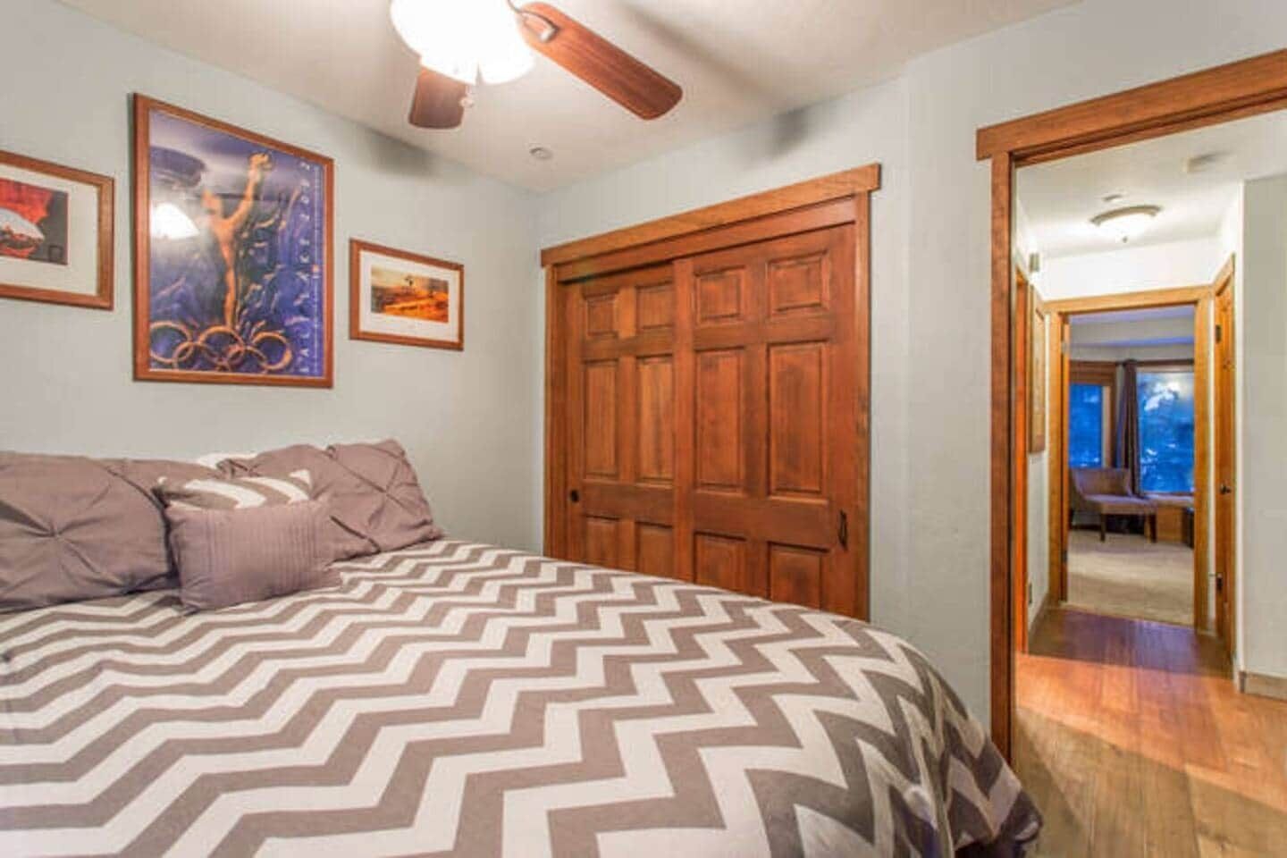 Een slaapkamer met een groot bed, een houten garderobe en een plafondventilator. Er hangen afbeeldingen in de muur en er is uitzicht op een andere kamer door een geopende deur.