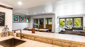 Een moderne keuken met een marmeren aanrecht, een spoelbak en een eethoek met een tafel en stoelen. Er zijn grote ramen met uitzicht op de omgeving.