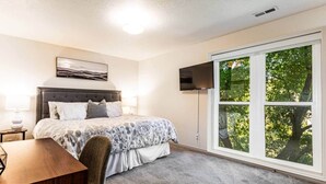 Een slaapkamer met een bed, een bureau, een stoel, een televisie en een groot raam met uitzicht op bomen.