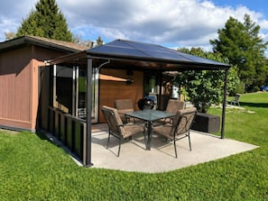 A covered patio with a table and chairs, a grill, and a view of a house.
