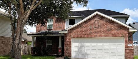 A two-story brick house with a white garage door, a covered front porch, and a tree in the front yard.