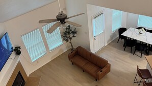 Ein geräumiger Wohnbereich mit Deckenventilator, einem braunen Sofa, einem Esstisch mit Stühlen und einem Fernseher an der linken Wand.