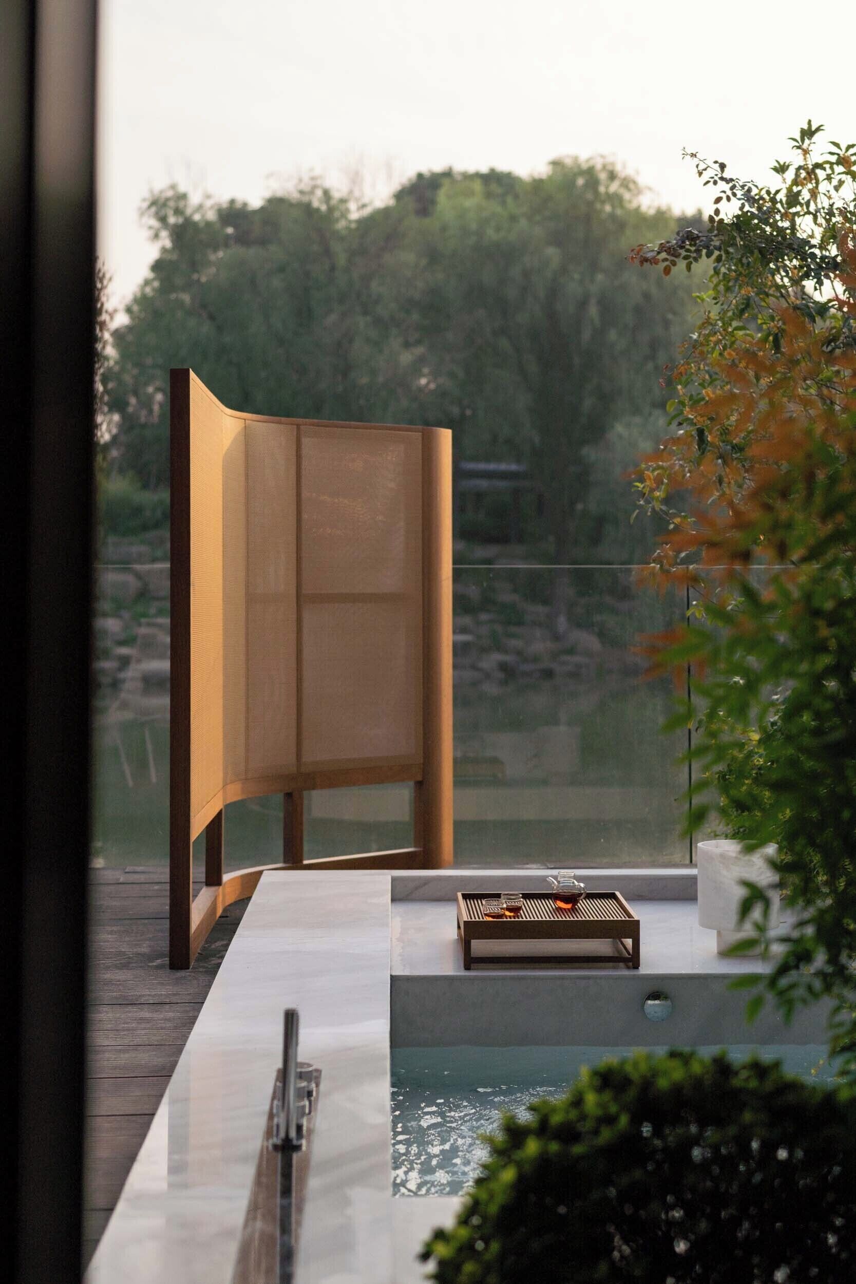 A modern outdoor pool area with a wooden deck, a seating area with a glass table, and a view of a natural landscape.