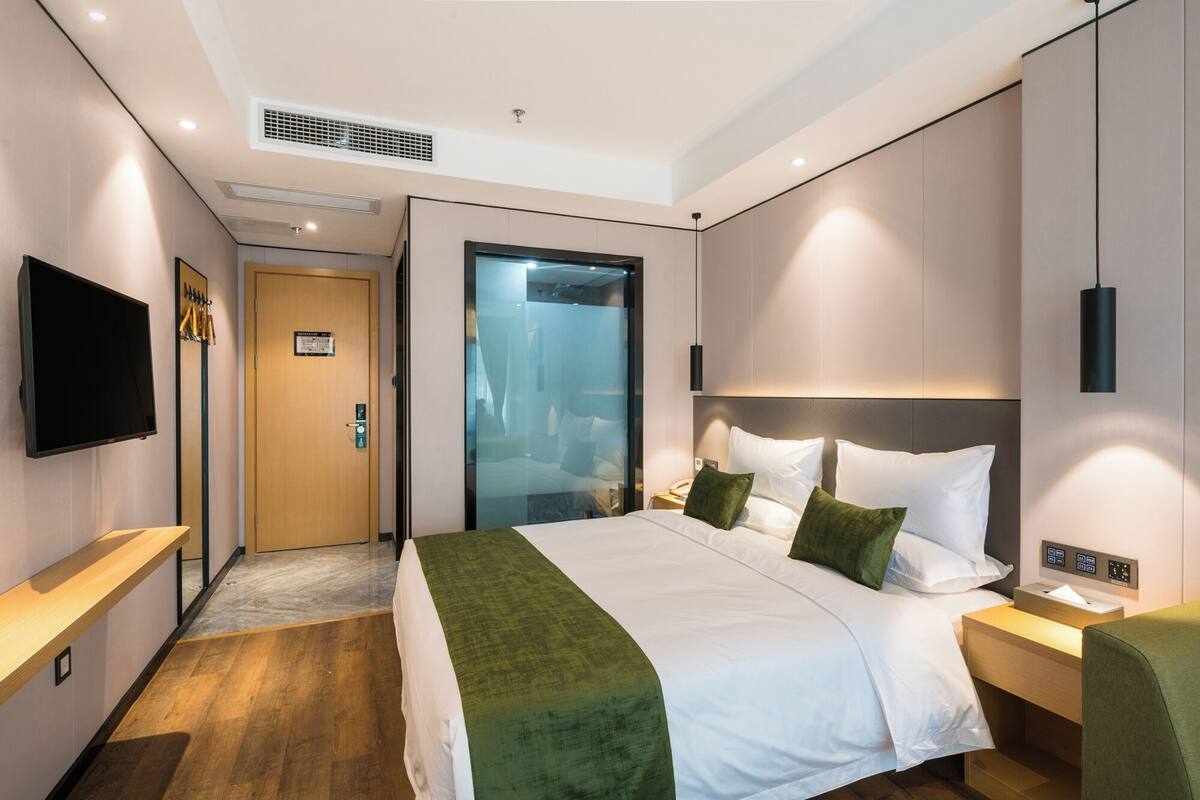 A modern hotel room with a large bed, a TV, a desk, and a glass door leading to a bathroom.