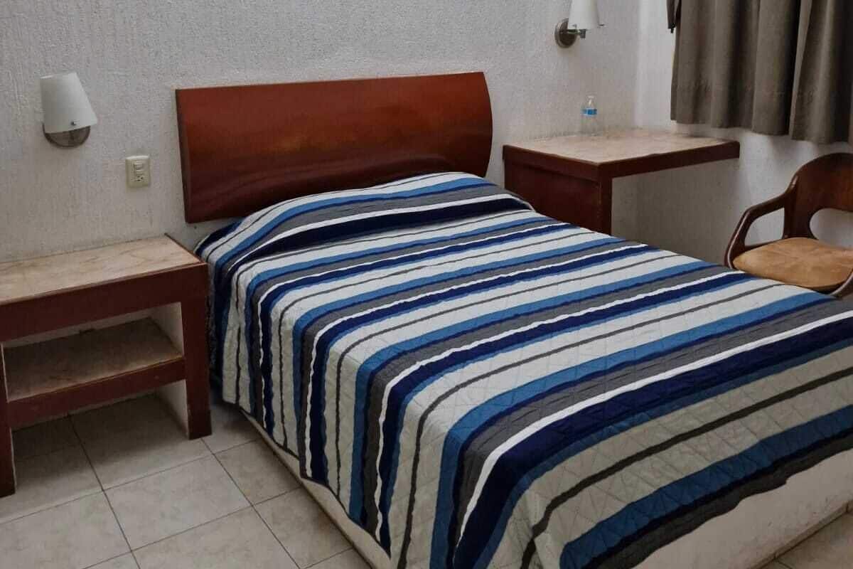 A single bed with a striped bedspread, two bedside tables, a nightstand, a lamp, a chair, and a curtain.