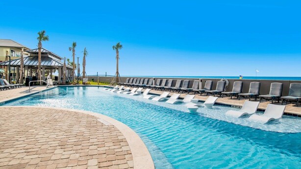 A pool area with lounge chairs and palm trees, a clear sky, and a view of the ocean. - The Oasis 1804 | Beautiful, spacious, beach front condo! Endless Amenities! (Orange Beach)