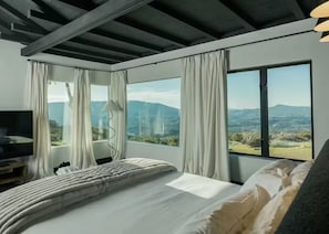 Quarto com uma cama grande, televisão e vista para montanhas através de amplas janelas.