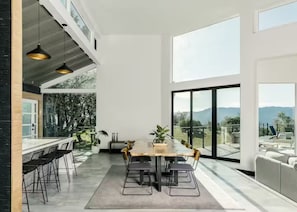 Ein moderner Essbereich mit einem langen Tisch und Stühlen, großen Fenstern mit Blick ins Freie und einem Balkon mit Sitzgelegenheit.