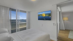 Een hotelkamer met een bed, een aan de muur bevestigde televisie, een balkon met uitzicht op zee en een deur die leidt naar een andere kamer.