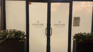 Una entrada doble con letrero 'VENTURE COMMONS', un felpudo metálico y plantas en macetas a ambos lados.