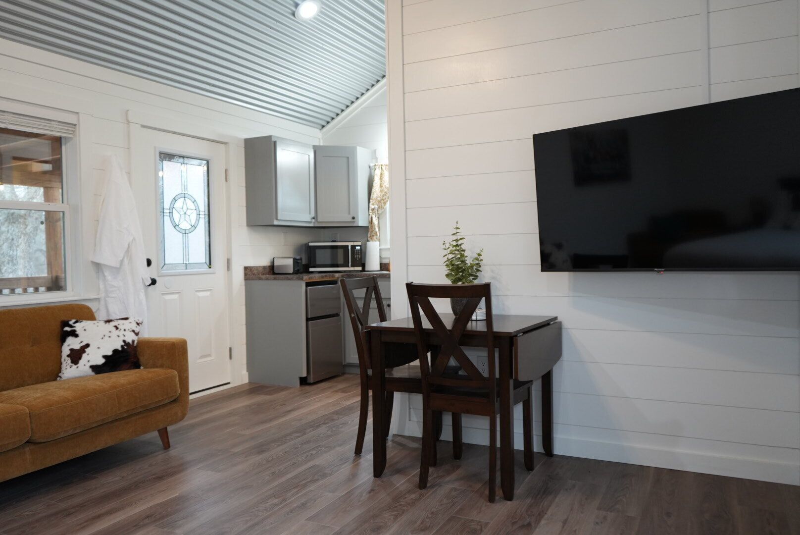 Une cuisine moderne avec des meubles blancs, un four à micro-ondes et un réfrigérateur. On y trouve un coin repas avec une table et des chaises en bois, ainsi qu’un téléviseur fixé au mur.