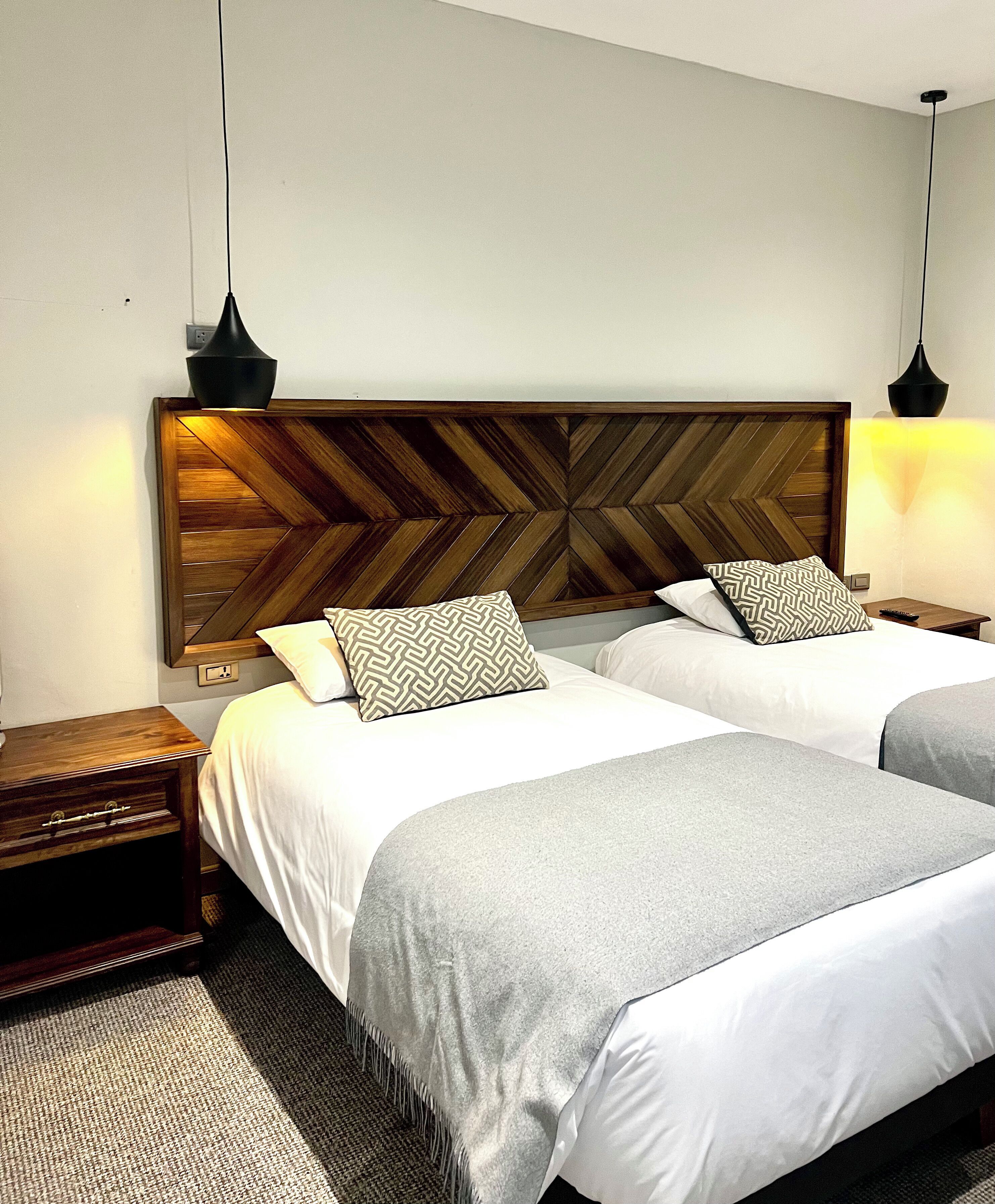 A hotel room with two beds, wooden headboards, bedside tables, and hanging lamps.