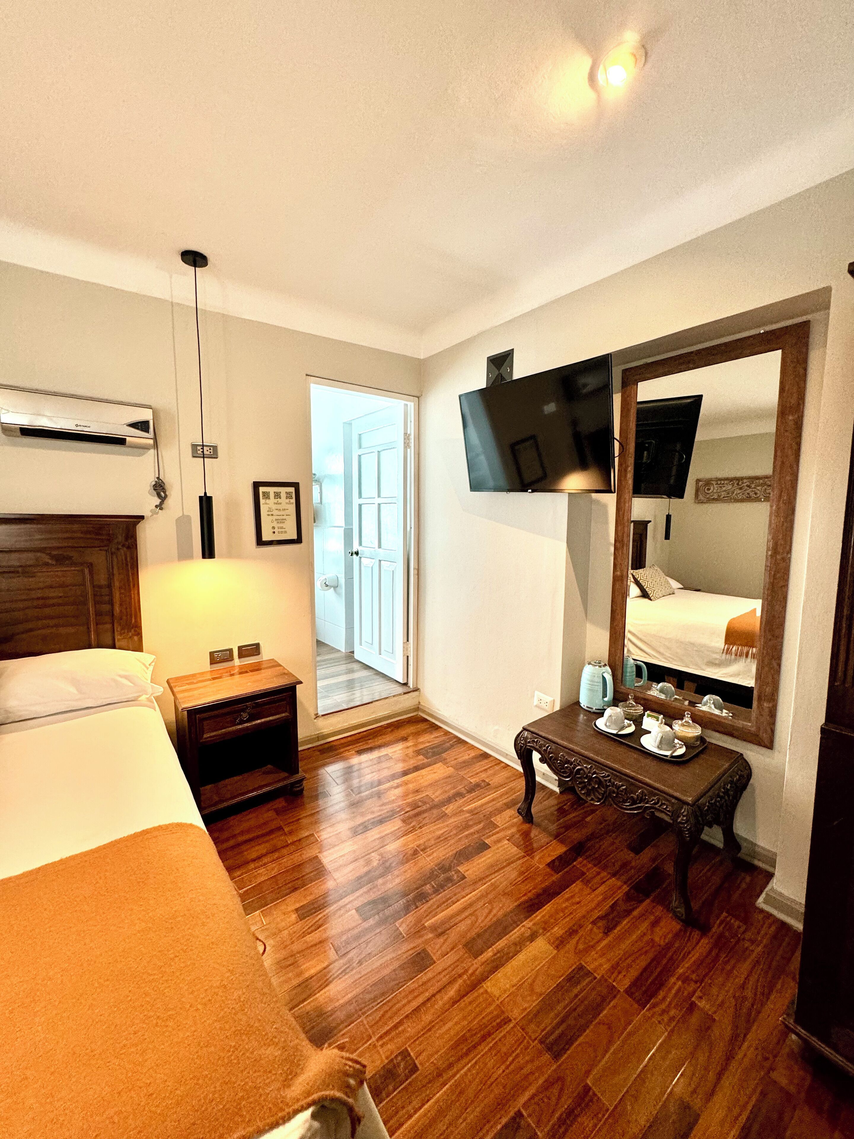 A hotel room with a bed, bedside table, dresser, wooden floor, and a large mirror.