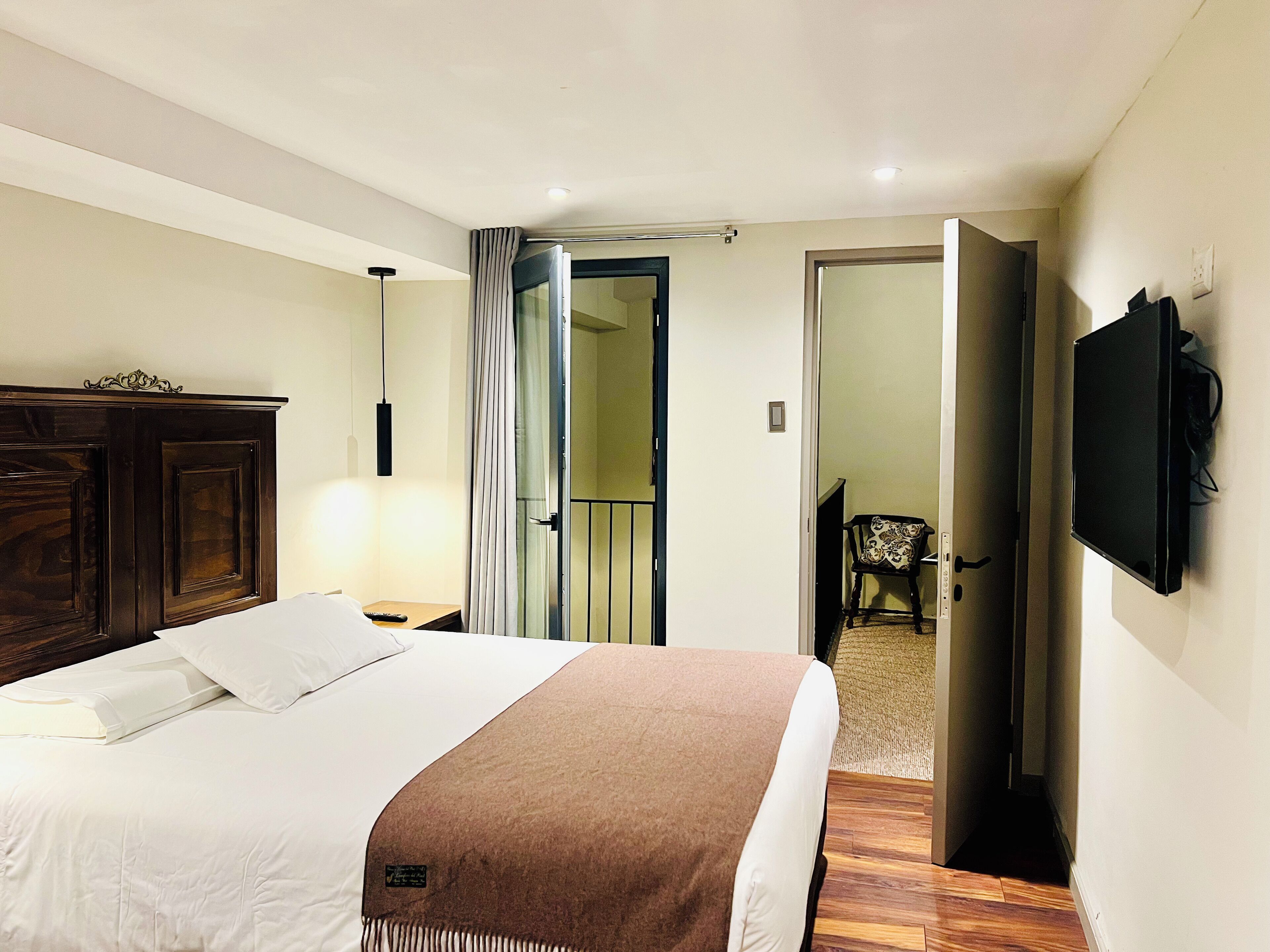 A hotel room with a bed, a wooden wardrobe, a TV mounted on the wall, and a door leading to another room.