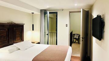A hotel room with a bed, a wooden wardrobe, a TV mounted on the wall, and a door leading to another room.