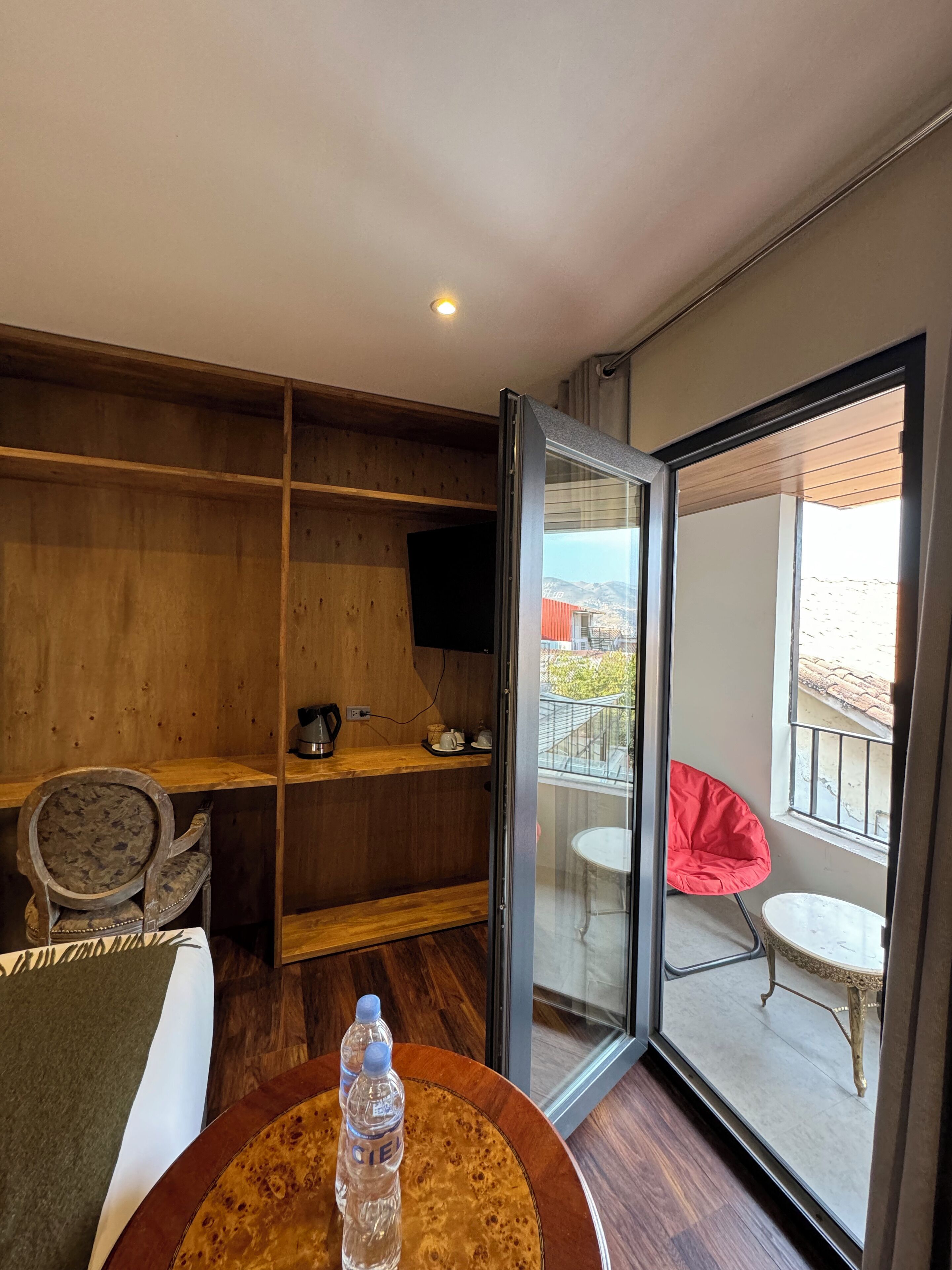 Una habitación de hotel moderna con un mueble de pared de madera, una cama individual, una mesa redonda con una botella, una silla roja y un balcón con vistas.
