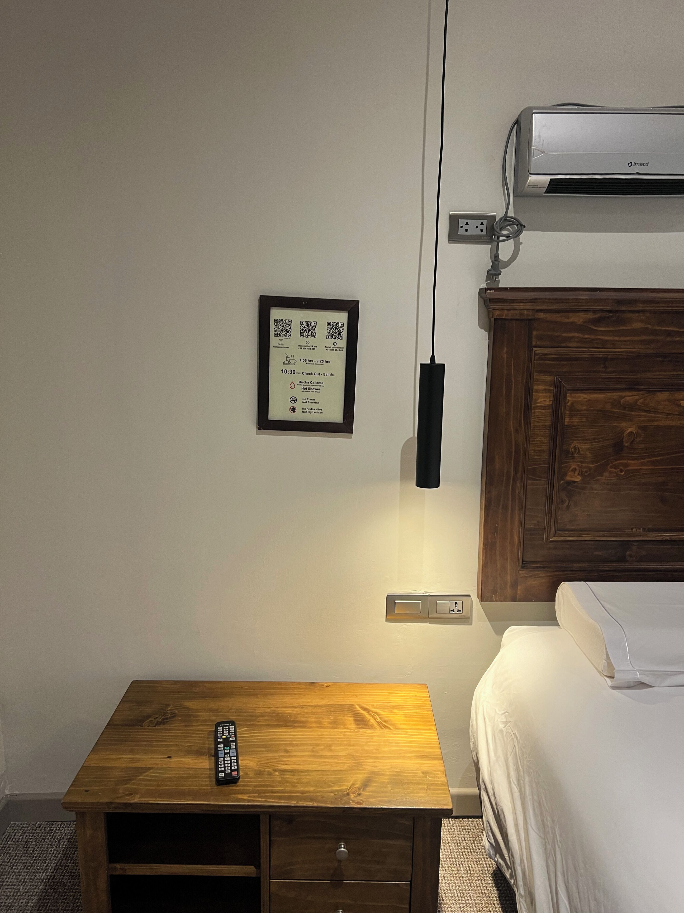 Una habitación de hotel con una mesita de noche de madera, una cama, un aire acondicionado instalado en la pared y un cartel con un código QR adherido a la pared.