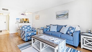 Un salon avec un canapé bleu, un fauteuil assorti et une table basse. On y trouve également un coin repas avec une table et des chaises, et une cuisine à l’arrière-plan.