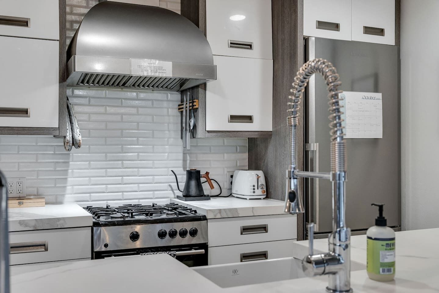 A modern kitchen with white cabinets, a stainless steel range hood, a gas stove, a kettle, a toaster, and a faucet with a soap dispenser.