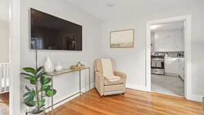 Un salón con un sillón color crema, un televisor de pantalla plana, una planta y una mesita. Se ve una cocina a través de un vano abierto.