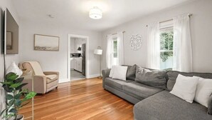Una sala de estar moderna con un sofá gris, un sillón color beige y suelo de madera. Se ve una cocina a través de un vano abierto.