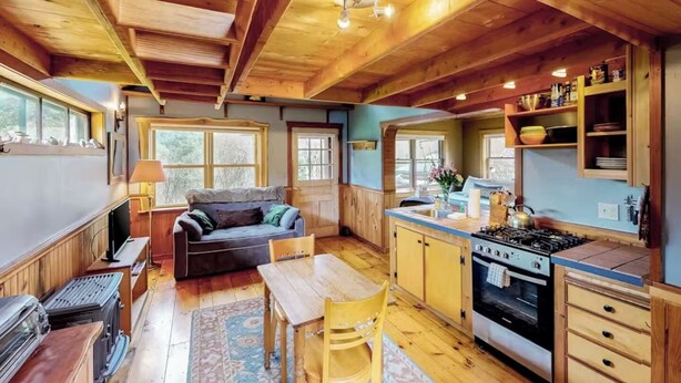 A cozy cabin interior with a wooden dining area, a sofa, a kitchen with a stove, and large windows. - VT Guest Cottage at Sugarbush in Moretown, VT (Northfield)