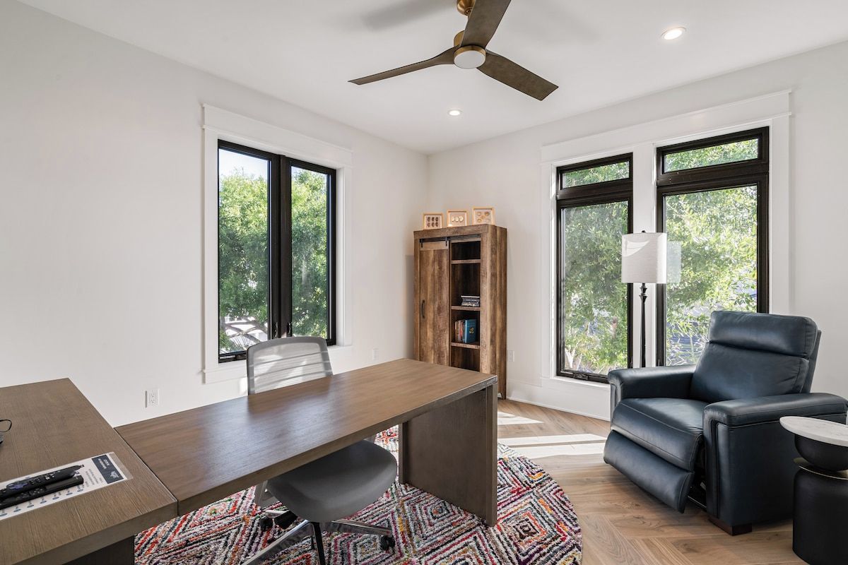 Una habitación moderna con escritorio, silla y sofá de cuero negro. Cuenta con ventilador de techo, estantería y ventanas amplias con vistas a árboles.