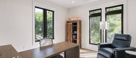 Una habitación moderna con escritorio, silla y sofá de cuero negro. Cuenta con ventilador de techo, estantería y ventanas amplias con vistas a árboles.