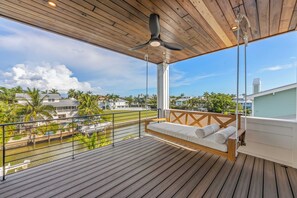 Une terrasse en bois avec un fauteuil suspendu, un ventilateur de plafond et une vue sur une rivière et des bâtiments.