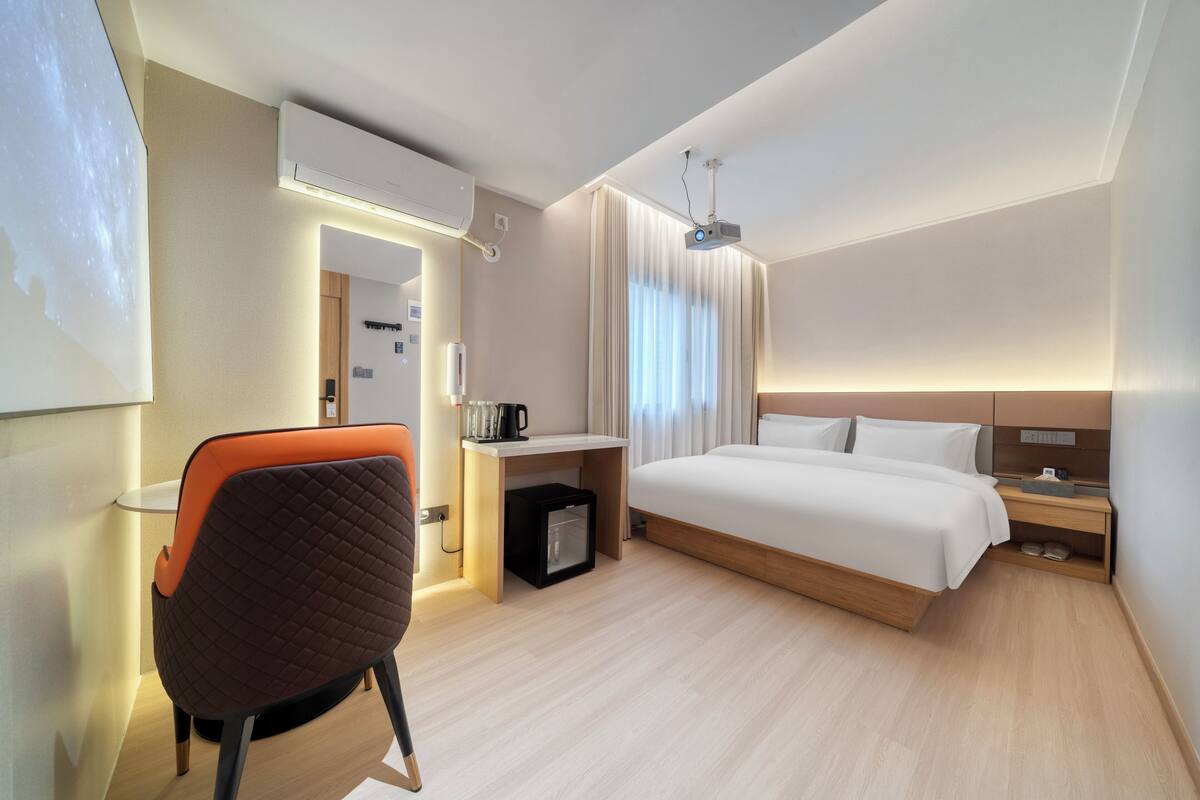 Una habitación de hotel moderna con cama, escritorio, silla, proyector y un mini refrigerador.
