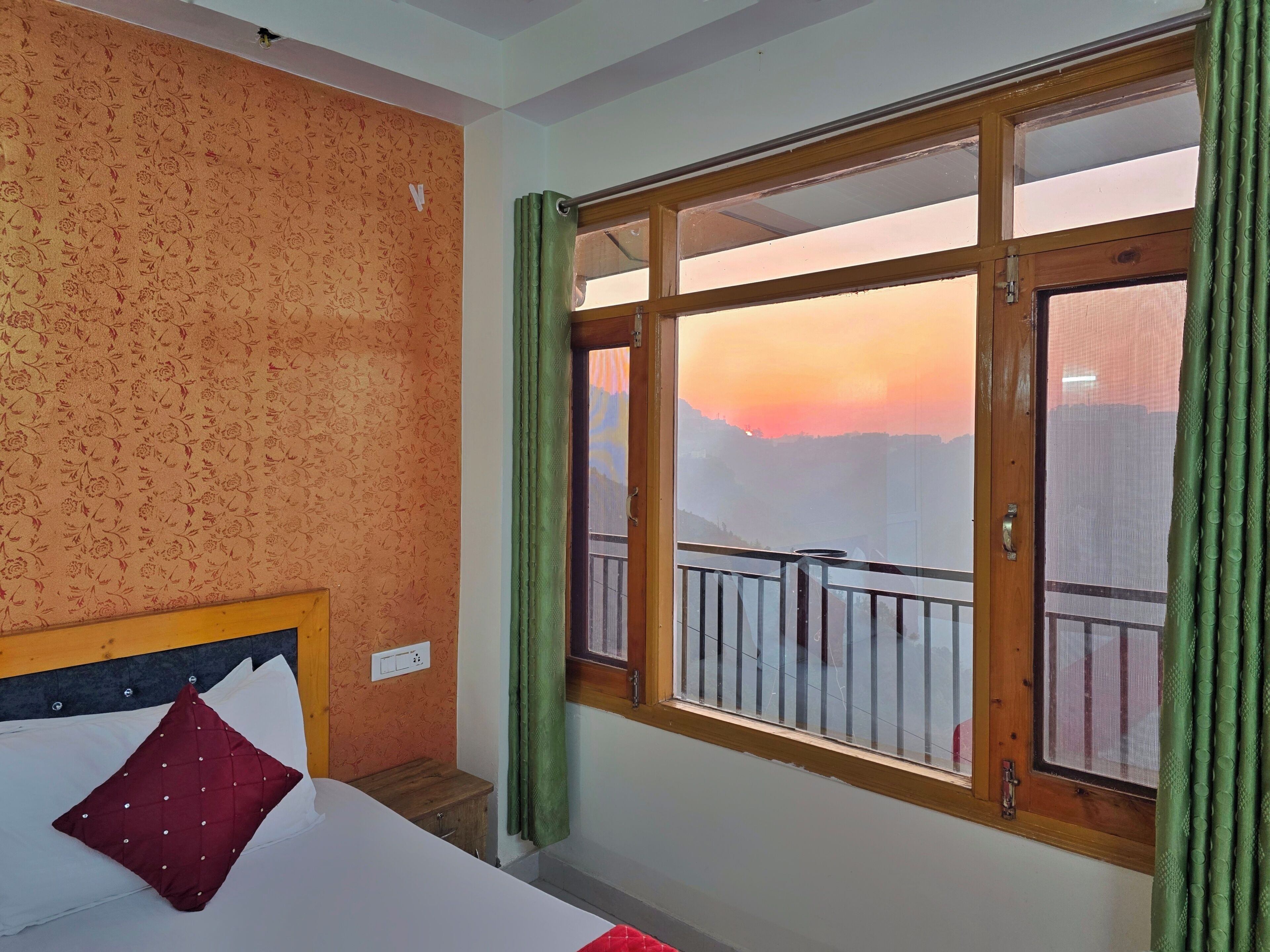Une chambre d’hôtel avec un lit, une petite table de chevet en bois, une fenêtre donnant sur un paysage urbain au coucher du soleil, et des rideaux verts.