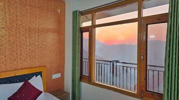 Une chambre d’hôtel avec un lit, une petite table de chevet en bois, une fenêtre donnant sur un paysage urbain au coucher du soleil, et des rideaux verts.