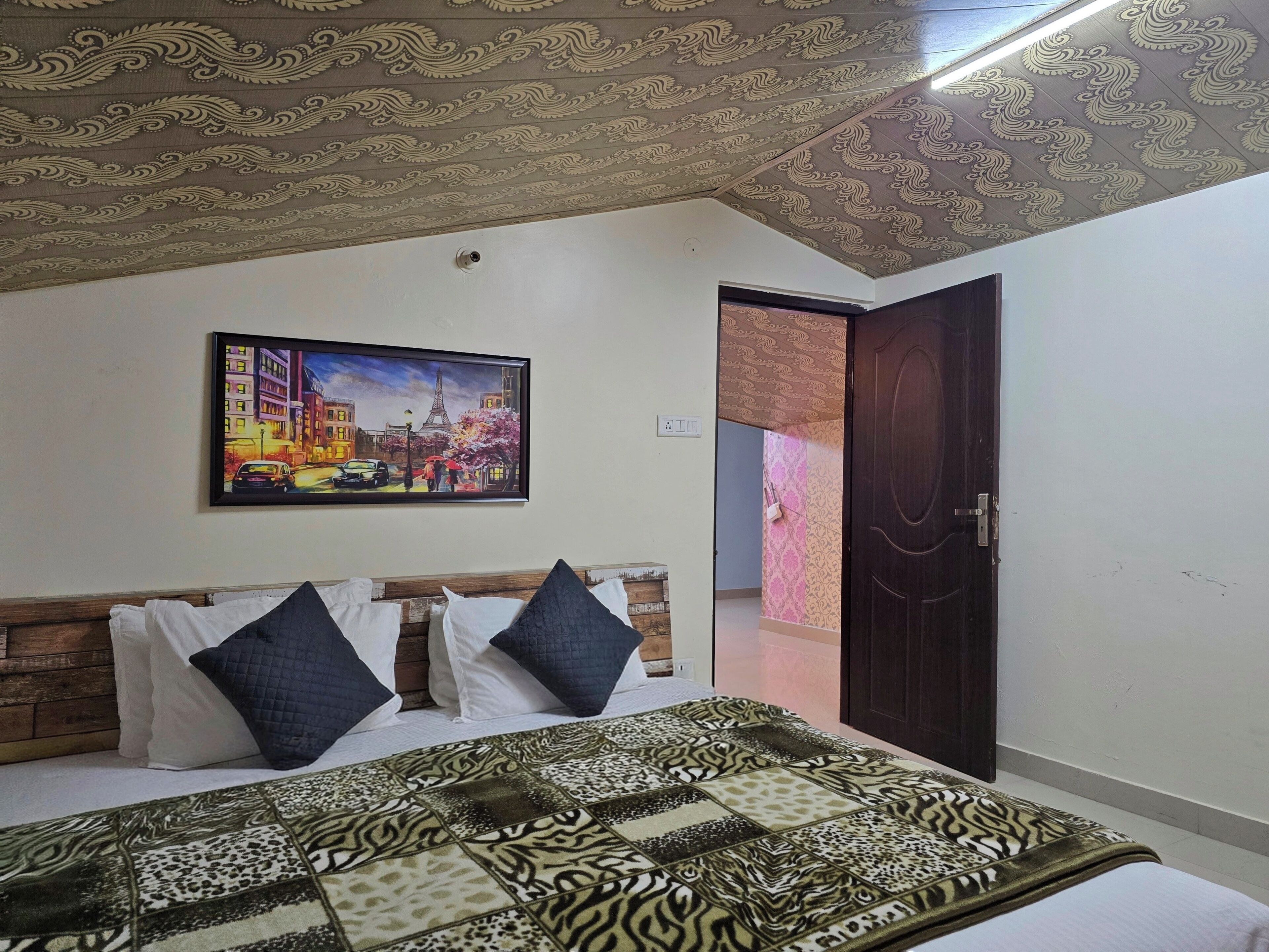 A bedroom with a patterned bedspread, a framed cityscape painting, and a door leading to another room.