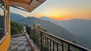 Uma varanda com chão xadrez, grade e vista para uma paisagem montanhosa ao pôr do sol.
