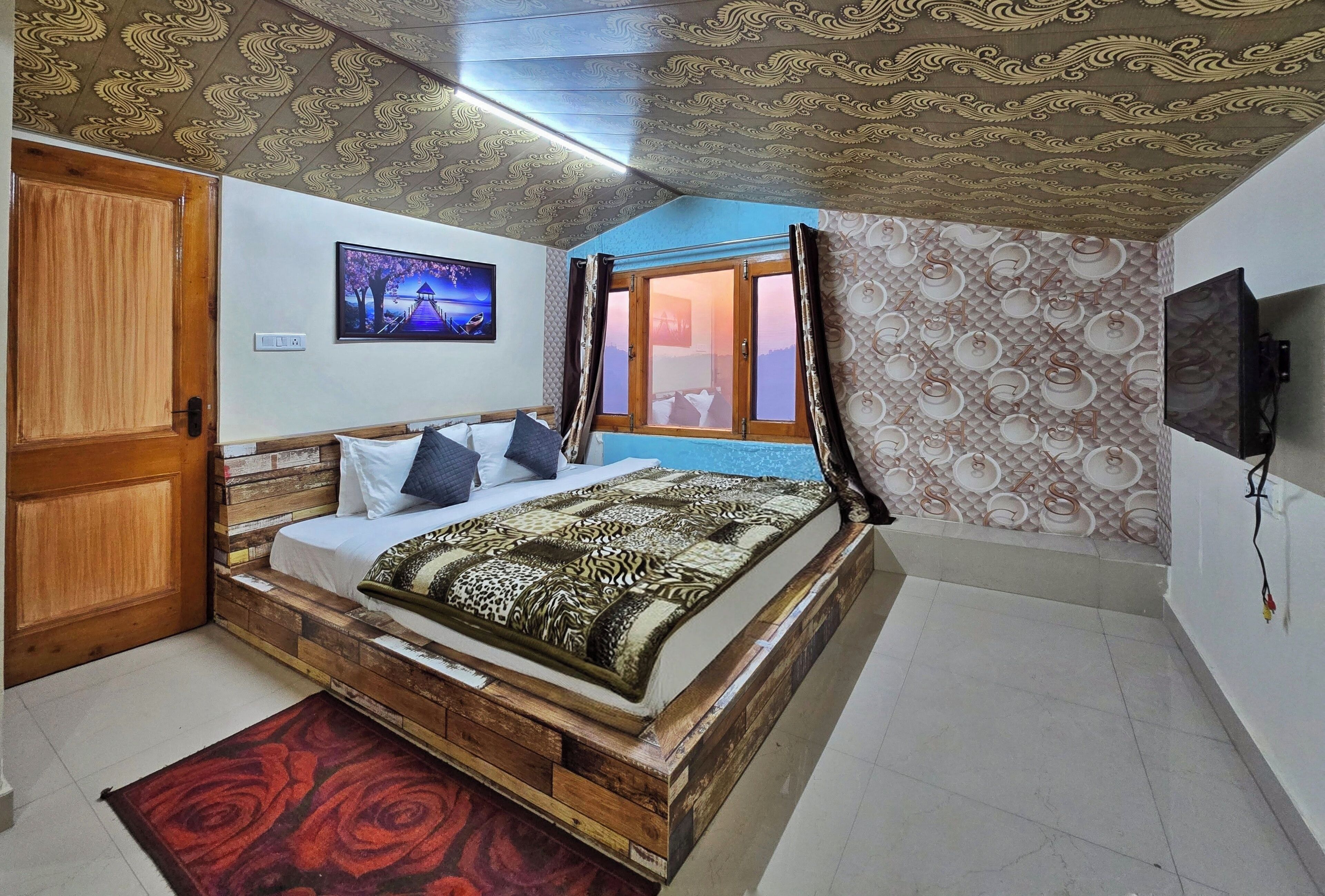 A bedroom with a bed, a wooden door, a window with curtains, a TV on the wall, and patterned wallpaper.