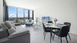 Un salon moderne avec un canapé, un fauteuil bleu, une table à manger dressée pour deux personnes et un grand écran de télévision.